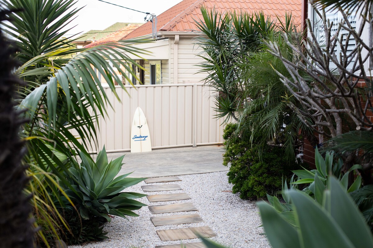A narrow pathway lined with tropical plants leads to a gated entrance. Soft gravel and stepping stones guide the way. A surfboard is propped against the fence, hinting at a coastal lifestyle.