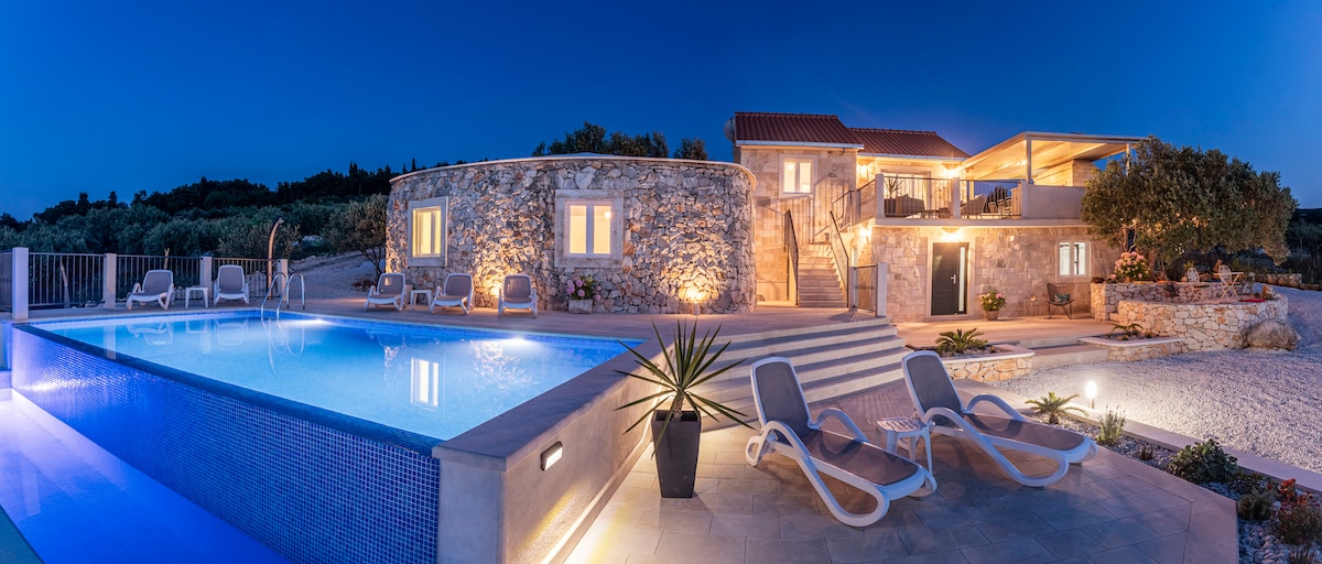 The villa's exterior is illuminated under a twilight sky, highlighting a spacious stone structure with warm lighting. A tranquil pool reflects the surrounding greenery and there are relaxing lounge chairs positioned nearby, creating an inviting outdoor area.