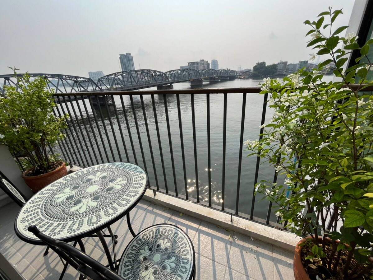A small balcony features a round patterned table and two chairs, surrounded by potted plants. View of the Chao Phraya River is visible, with shimmering reflections on the water and a distant cityscape, enhancing the tranquil outdoor setting.