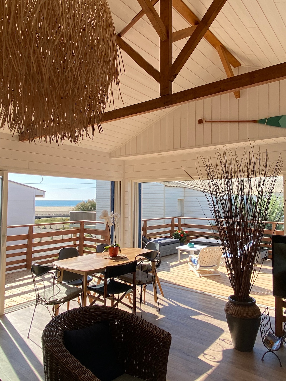 A bright living area features large open doors leading to a spacious terrace. A dining table with chairs stands alongside a comfortable seating area. Decorative elements include a large pendant light, potted plants, and tall decorative branches in a vase.