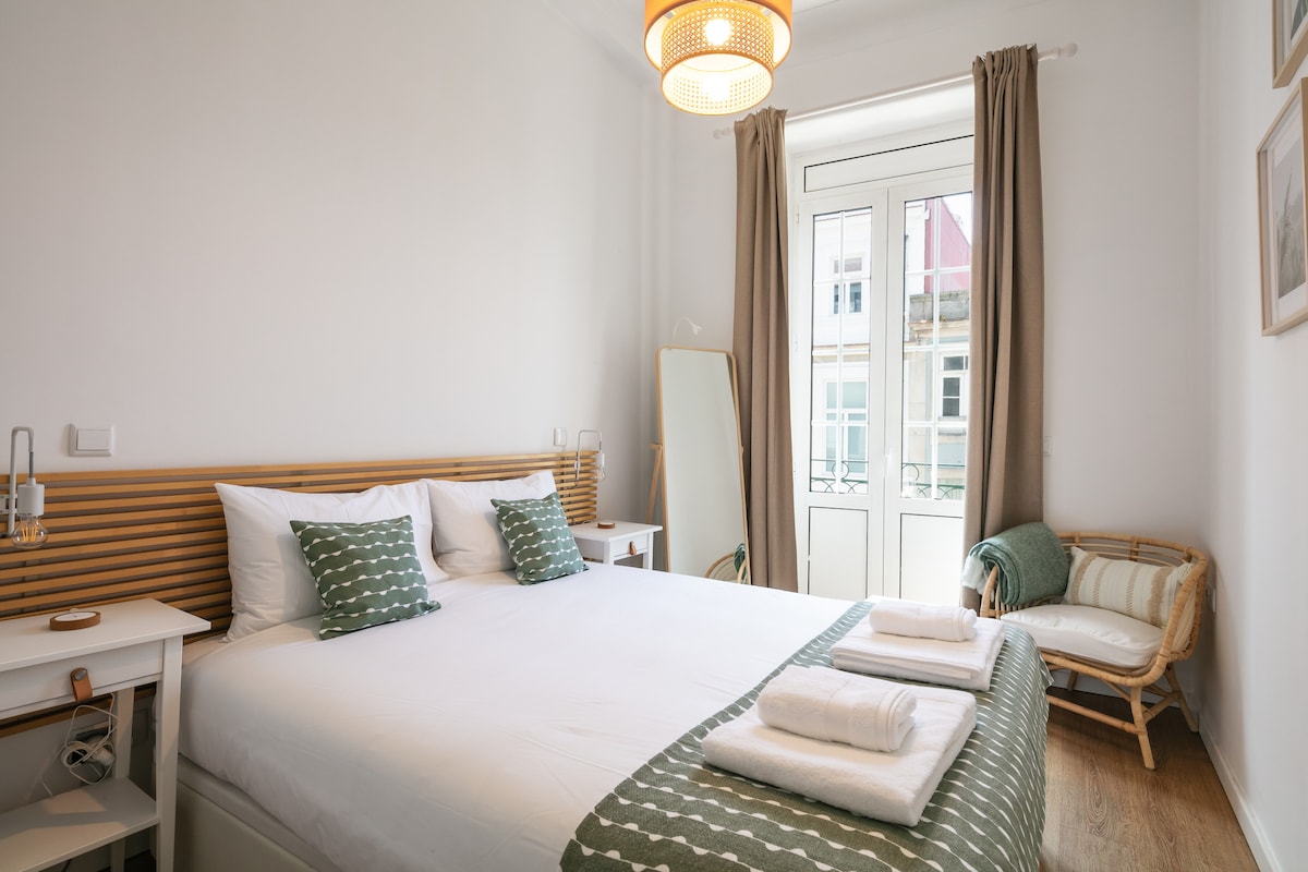A bright and airy bedroom features a spacious bed adorned with white linens and patterned throw pillows. Natural light floods the room through a large window framed by light curtains. A complementary chair and a mirror enhance the functional layout.