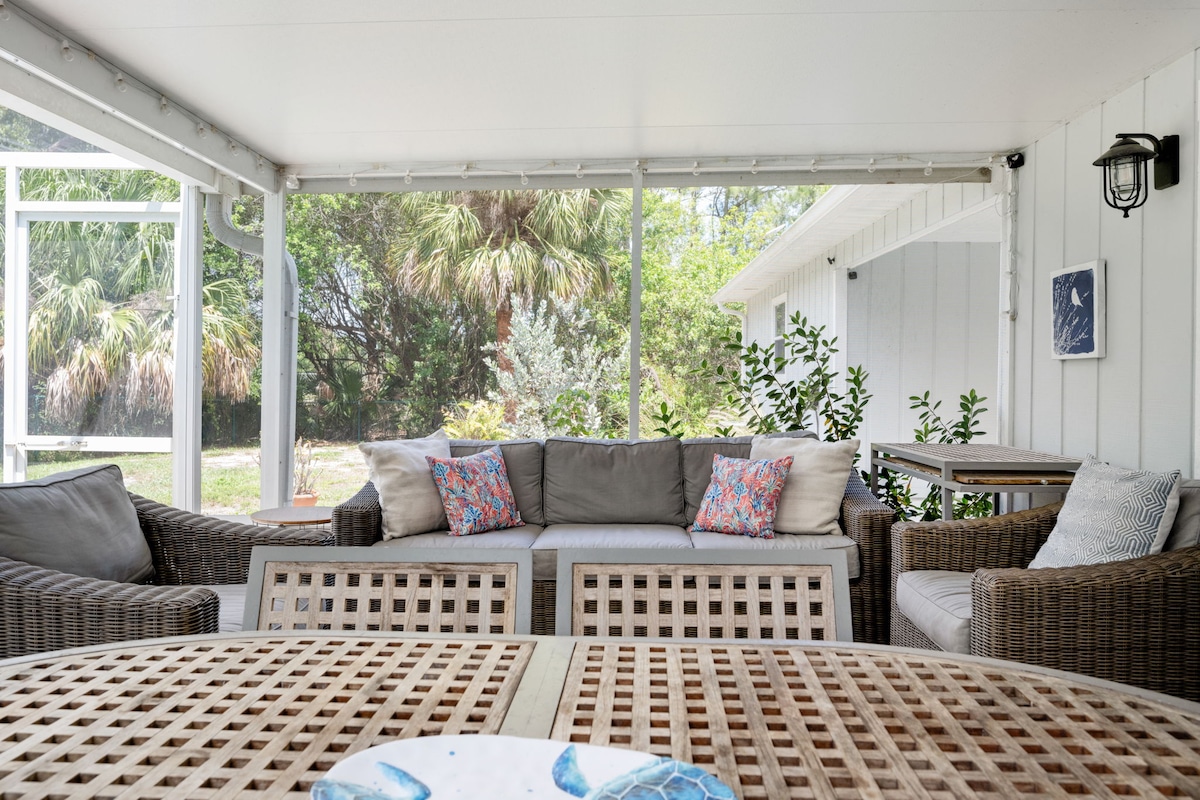 An inviting lanai area features comfortable seating arranged around a coffee table, with cushions in soft hues. Natural light filters through screens, illuminating surrounding plants and trees that enhance the serene outdoor environment.