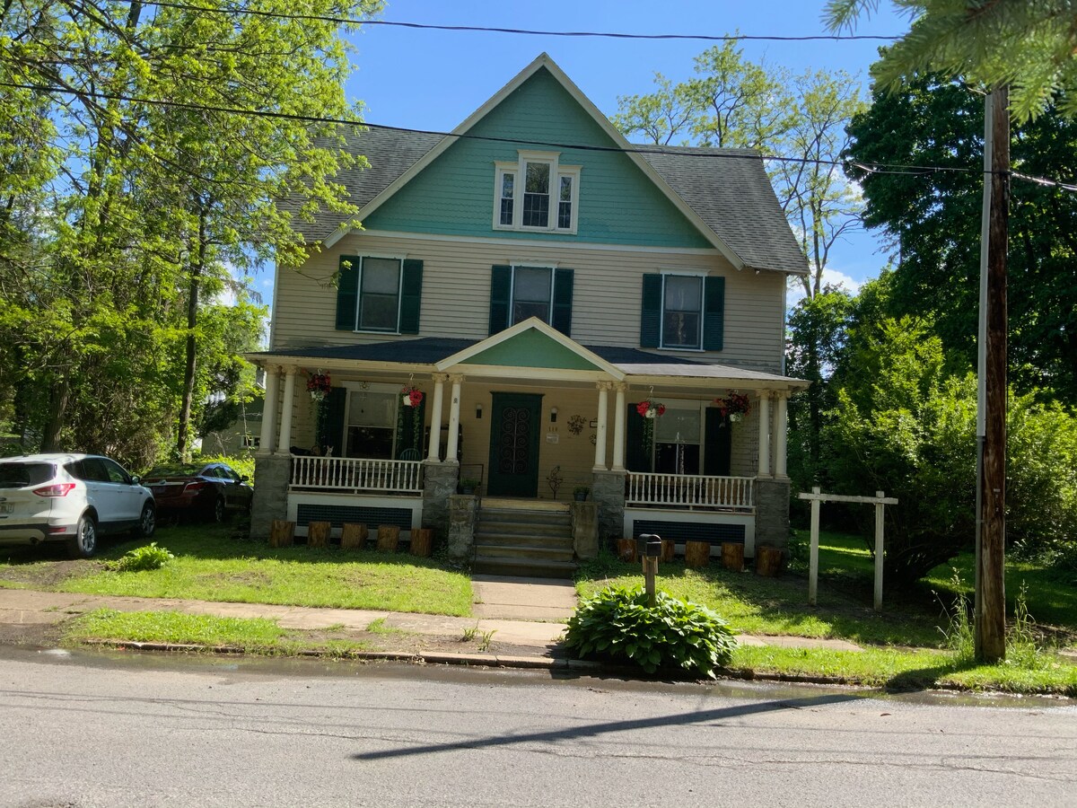 A charming house features a pale blue exterior with a welcoming porch. Hanging flower baskets adorn the front, and steps lead up to the main entrance. Lush greenery surrounds the property, providing a serene setting. A driveway is visible, accommodating parked cars.
