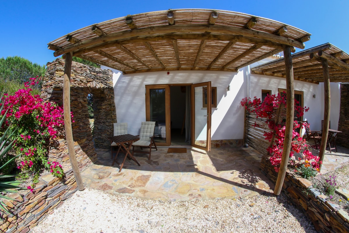 A charming outdoor patio is featured, surrounded by vibrant bougainvillea flowers. The stone pathway leads to a pair of wooden doors that open into the interior. Two cushioned chairs and a small table are positioned under a wooden pergola, providing a shaded area for relaxation.