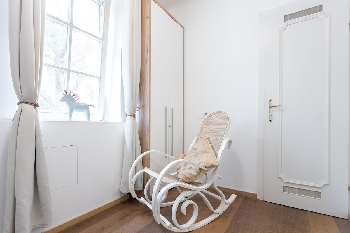 A cozy corner features a white rocking chair with a cushioned pillow, positioned beside a window. Gentle light filters through sheer curtains, and a minimalist wardrobe stands against the wall, contributing to a serene atmosphere.