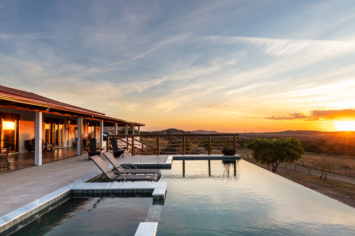 A modern house with expansive outdoor seating overlooks a tranquil pool featuring an infinity edge. The setting sun casts warm tones across the landscape, creating a serene backdrop. Lounge chairs are positioned beside the pool, inviting relaxation in a natural environment.