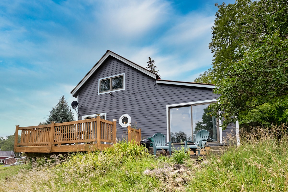 The exterior of the home is shown, featuring a deck with wooden railings and seating for outdoor relaxation. Large windows allow natural light to brighten the interior, and the surrounding landscape includes greenery and a clear blue sky, enhancing the serene environment of the property.