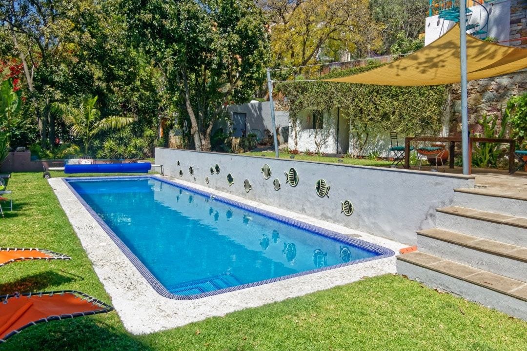 A spacious salt-water swimming pool is set within a lush garden, surrounded by palm trees and vibrant greenery. Sun loungers are arranged along the grass, and a shaded seating area is visible nearby, offering a comfortable spot to relax.