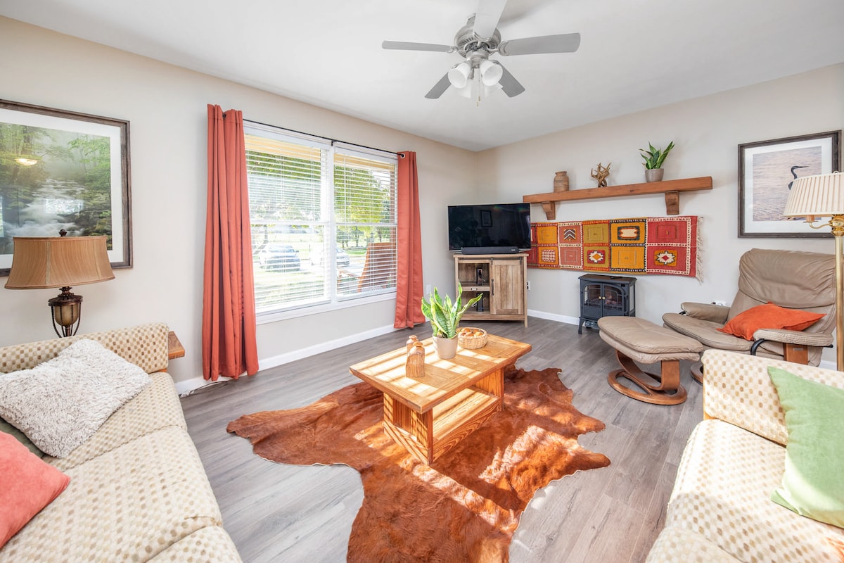 The living room showcases a cozy seating arrangement with a patterned couch and armchair. A wooden coffee table is placed on a textured rug, and a wall-mounted television is visible. Natural light enters through a large window, framed by orange curtains.