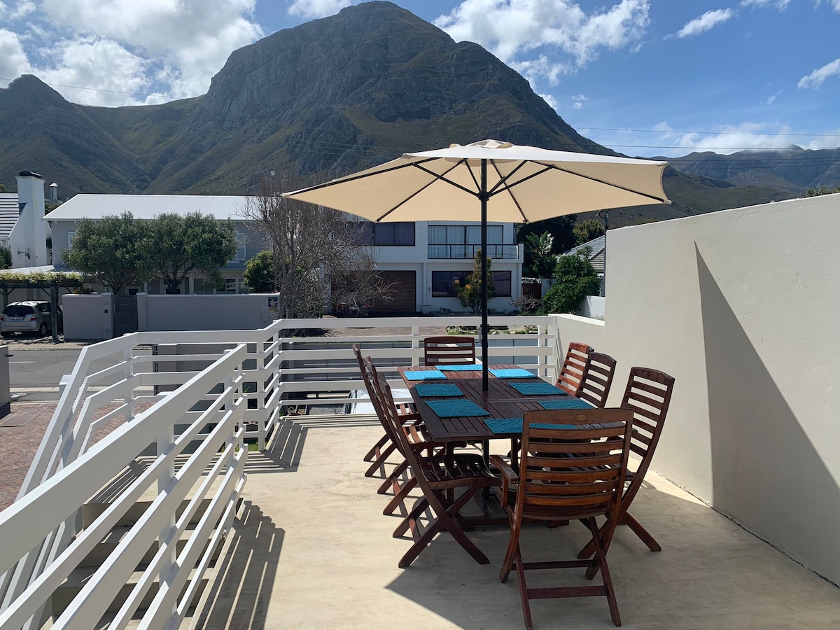 A spacious outdoor patio is furnished with a large wooden table and chairs, shaded by a large umbrella. The backdrop features a scenic mountain view under a clear blue sky, creating an inviting space for meals and relaxation.