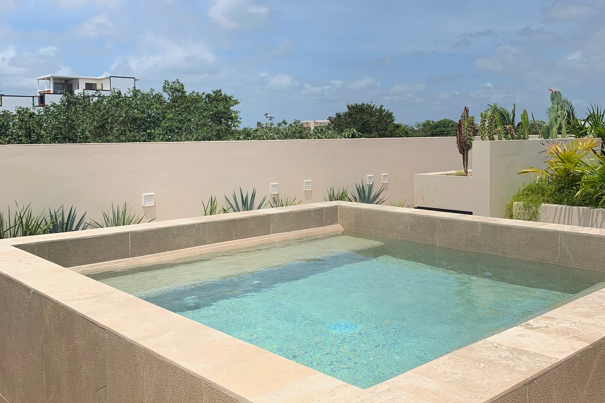 A tranquil pool area is depicted, featuring a square-shaped pool with clear water reflecting the sky. Surrounding greenery and minimalistic landscaping enhance the serene environment, while the smooth stone edges provide a seamless transition between the pool and the surrounding space.
