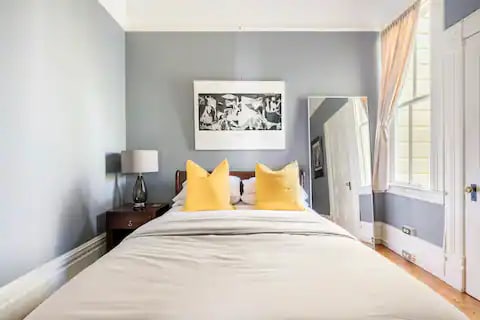 A private bedroom features a queen bed dressed in light-colored linens, complemented by oversized yellow pillows. A lamp and a side table sit next to the bed. A large mirror reflects the natural light from the window, enhancing the spacious feel of the room.