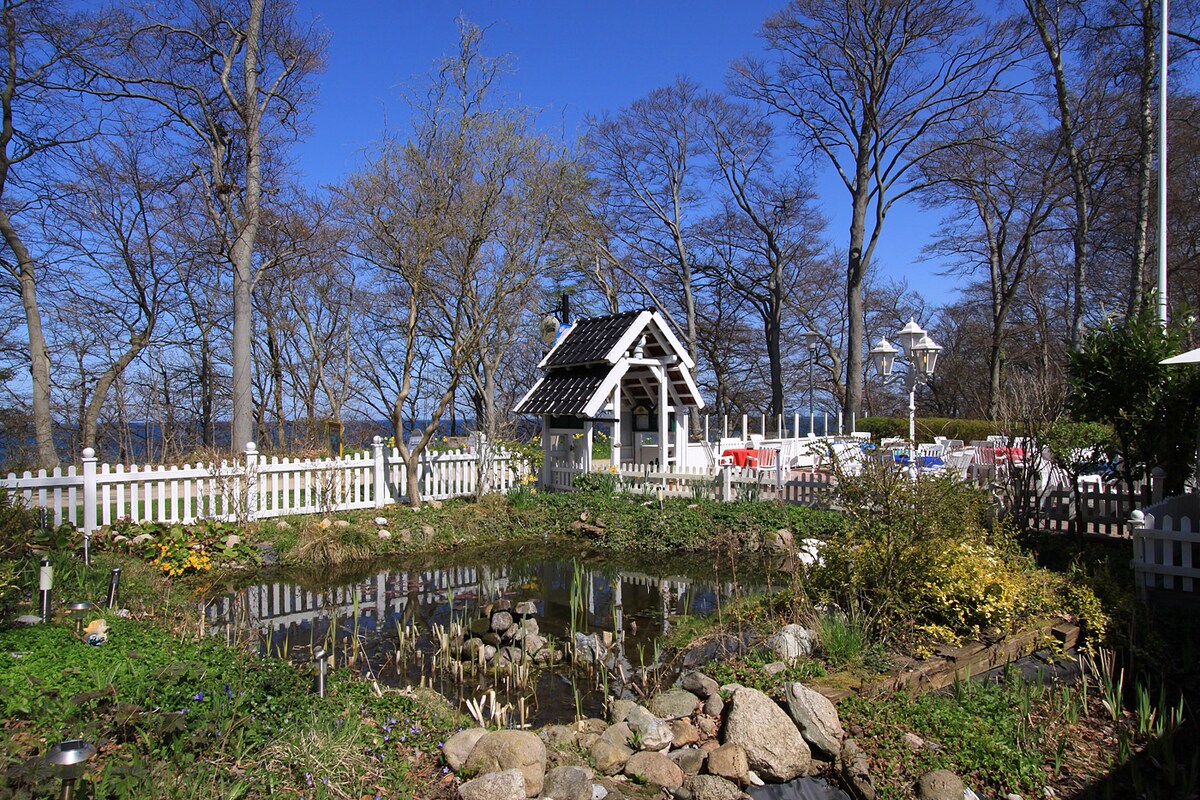 A tranquil garden area features a circular pond surrounded by stones and lush greenery. A small gazebo with a black and white roof stands nearby, along with white benches and tables set among trees that provide partial shade. Clear blue skies create a serene atmosphere.