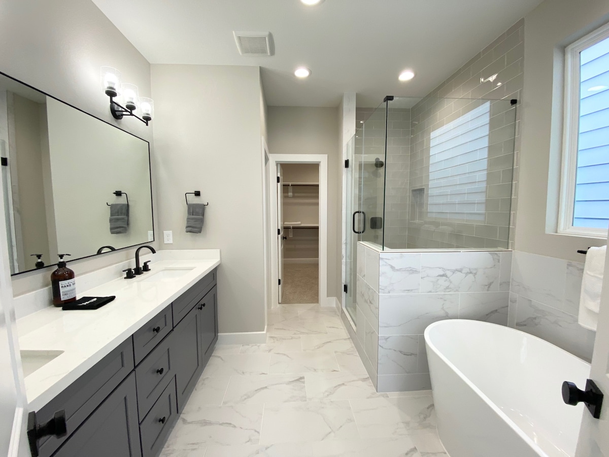 The bathroom features a spacious layout with double sinks set in a dark cabinet. A freestanding soaking tub is positioned beside a glass-enclosed shower. Natural light enters through a nearby window, complementing the neutral tile and marble accents throughout the space.