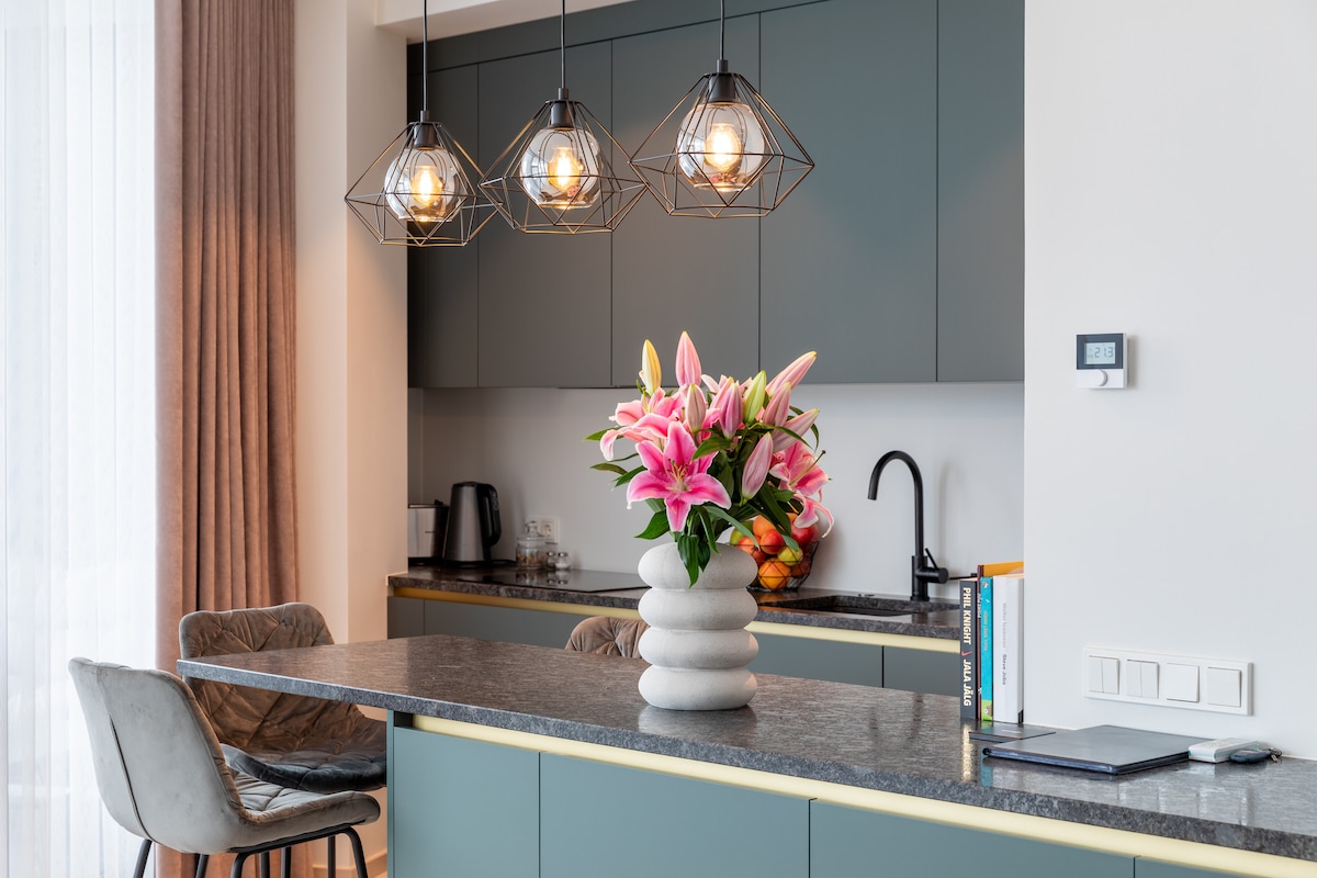 A modern kitchen is highlighted, featuring a dark stone countertop and stylish cabinetry in muted green. Three geometric pendant lights hang above, casting warm light. A vase with pink lilies adds a vibrant touch, while a coffee maker and fruit bowl are positioned on the counter.