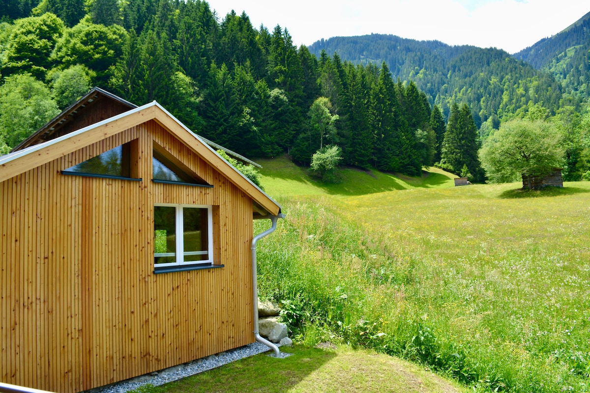 A small wooden structure is positioned amidst lush greenery, showcasing a sloped roof and large windows. Surrounding hills are visible in the background, covered with trees and patches of open grass, creating a peaceful natural setting.