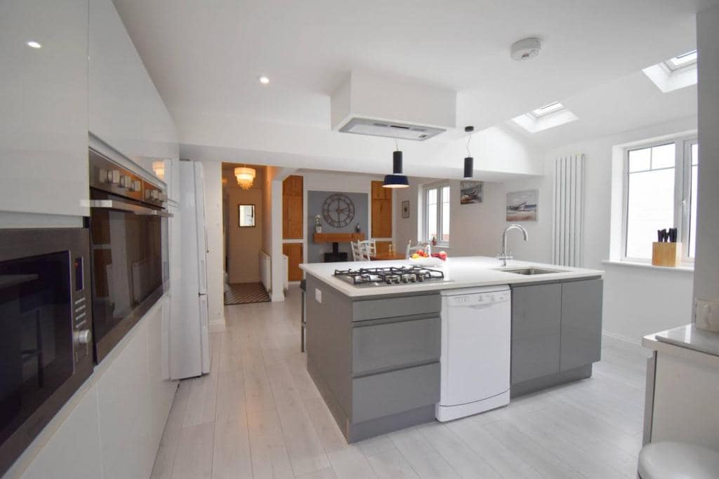 The open plan kitchen features a central island with a gas cooktop and a dishwasher. Modern cabinetry complements the sleek appliances, while natural light enters through multiple skylights and windows. A dining area is visible in the background, enhancing the spacious layout.