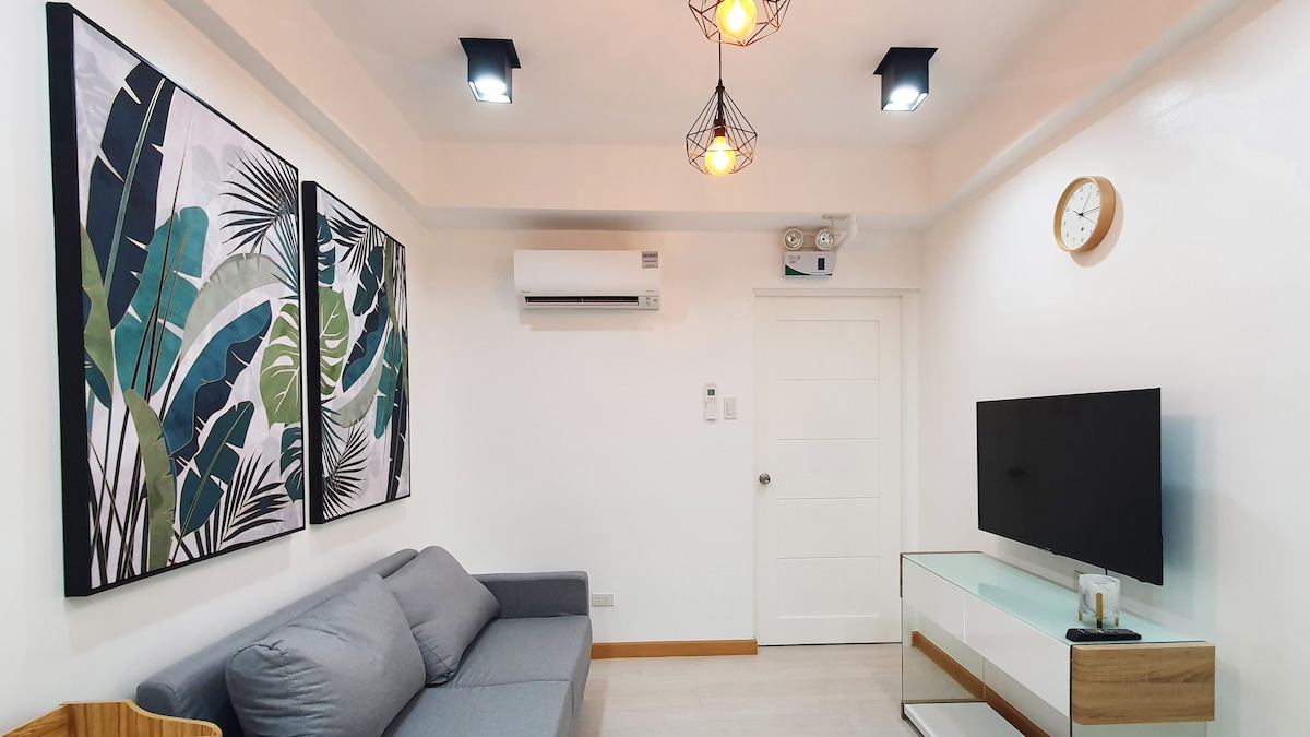 A cozy living space features a gray sofa positioned against the wall. Two large framed artworks with leafy designs are displayed, alongside a mounted flat-screen TV and a white console table. Ceiling lights provide bright illumination, while an air conditioning unit is installed on the wall.