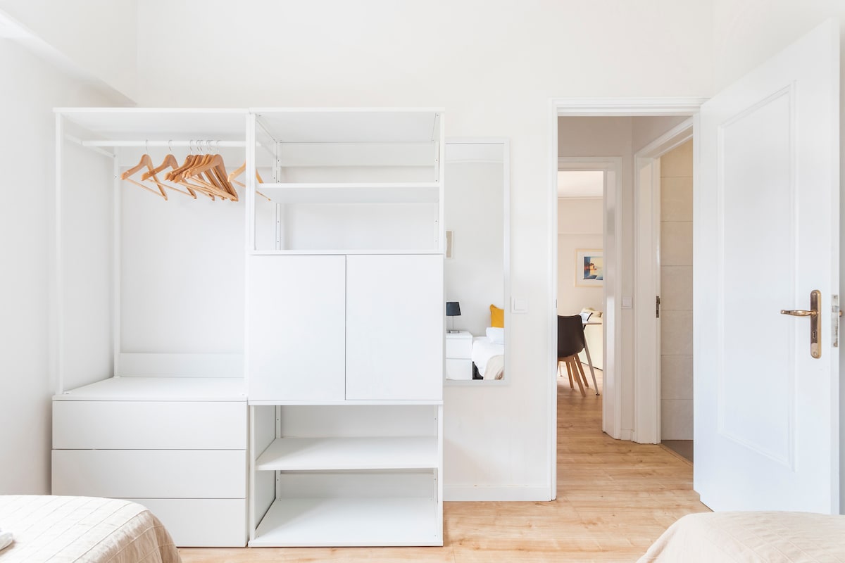 A bright bedroom features a built-in wardrobe with hanging space and drawers. Light wooden flooring runs throughout the room. A doorway leads to another area, revealing soft lighting that hints at a welcoming atmosphere.
