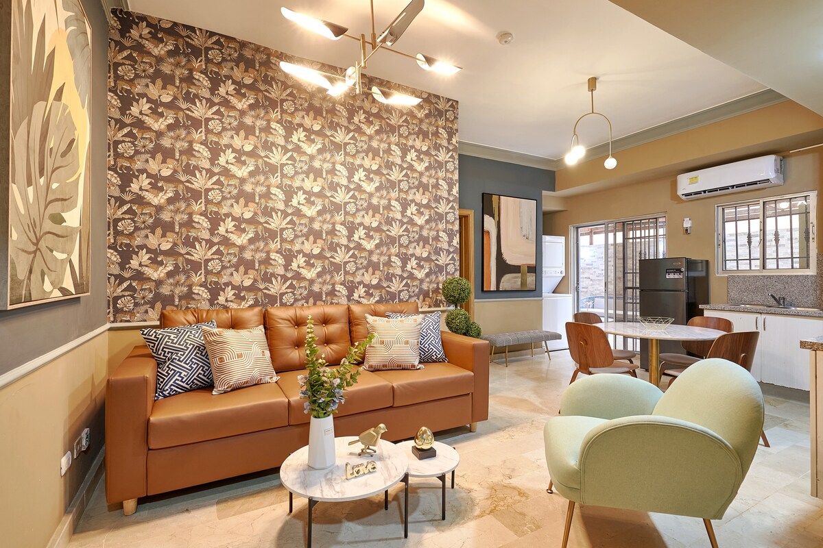 A well-furnished living area features a brown leather sofa adorned with decorative pillows, a round coffee table, and stylish accent chairs. A floral wallpaper adds texture, while natural light filters through the windows, highlighting the space's modern design.