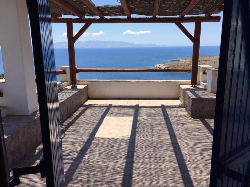 An outdoor terrace is framed by a wooden pergola, offering sweeping views of the sea and nearby islands. The sun casts playful shadows on the tiled floor. Clear skies and gentle waves create a serene backdrop, inviting relaxation in a scenic setting.