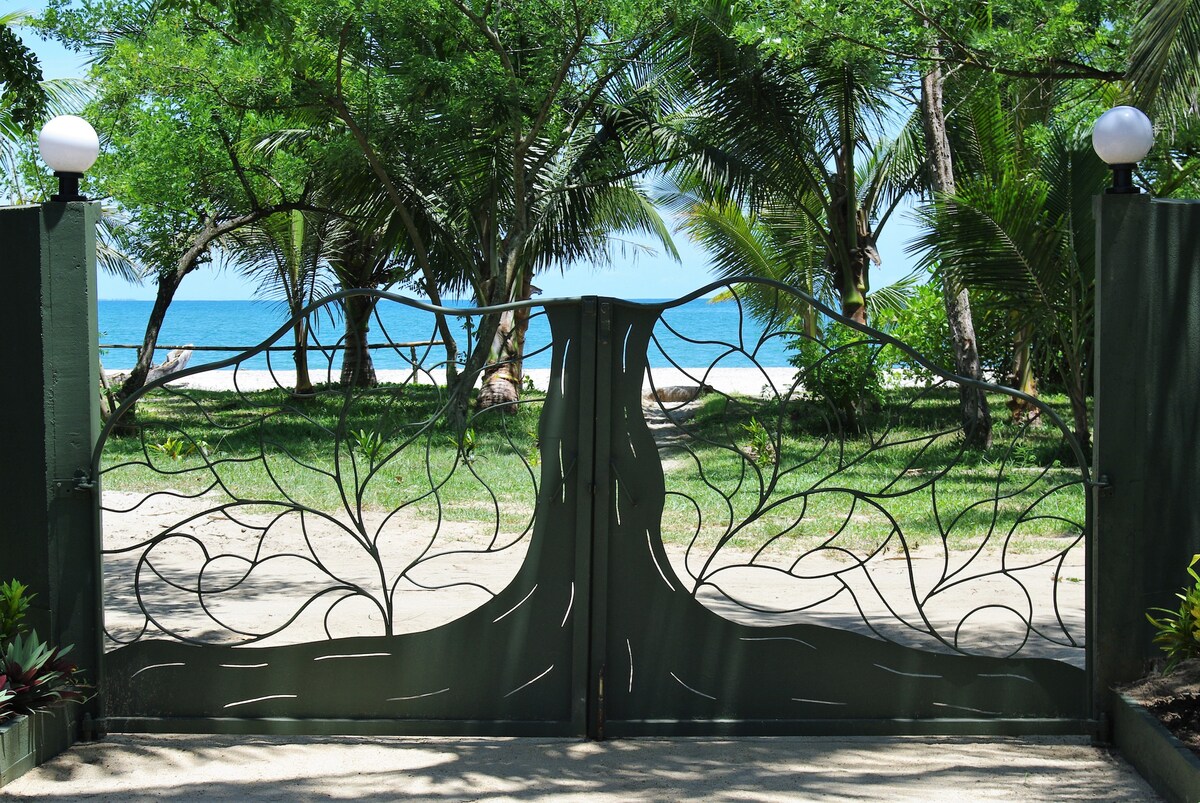 A decorative entrance gate crafted from intricate metalwork provides access to a sandy beach. Lush green palm trees frame the path, leading to the tranquil blue waters beyond. The scene reflects a serene coastal environment, inviting exploration of the nearby beach.