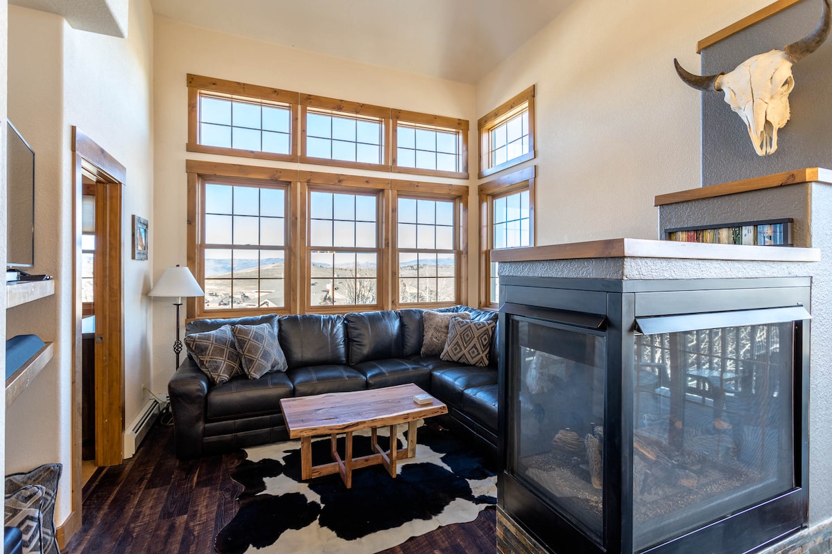 The living room features a spacious sectional sofa facing a gas fireplace. Large double high windows provide ample natural light and showcase mountain views. A wooden coffee table sits in the center, complemented by a decorative cowhide rug. A wall-mounted shelf holds books and decorations.