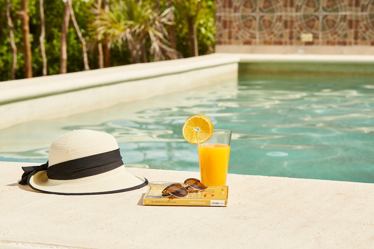 A sun hat rests on the stone edge of a tranquil pool, accompanied by a refreshing drink garnished with an orange slice. A book lies open beside the hat, creating a relaxed atmosphere in this inviting outdoor space.
