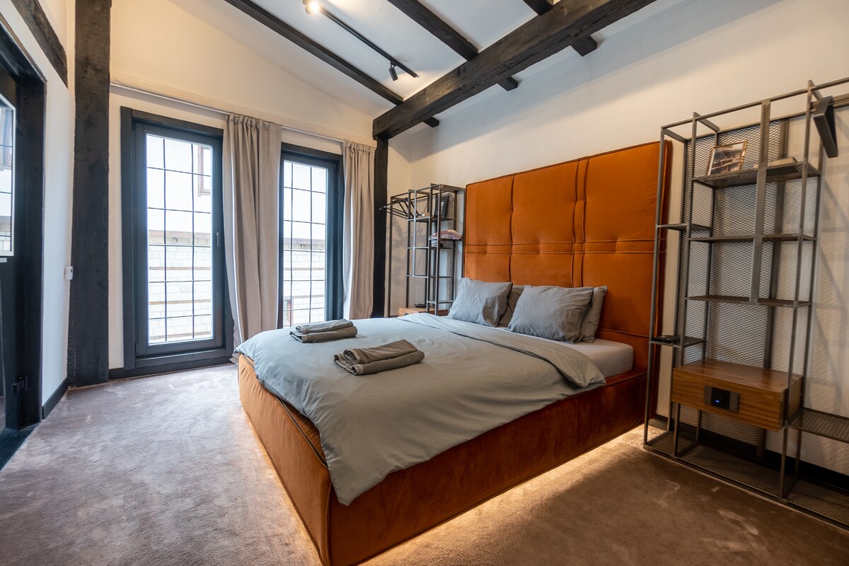 A spacious bedroom features a large orange upholstered bed with gray bedding. Two windows with black frames allow natural light to enter, while gray curtains hang beside them. A metal shelving unit adds functional storage, complementing the wooden beam accents on the ceiling.