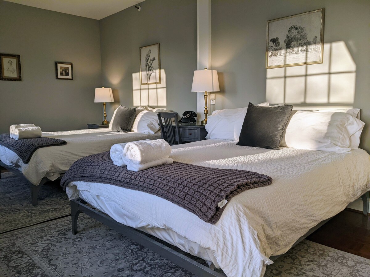 The guest room features two queen beds with textured throw blankets, neatly arranged at the foot of each bed. Soft light filters through the window, casting gentle shadows on the walls. A pair of bedside lamps add a touch of warmth to the cozy setting.