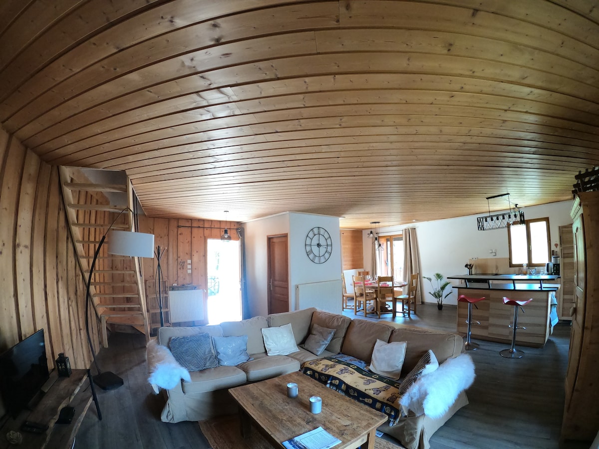A spacious living area features wooden paneling and a high ceiling, complemented by a large sectional sofa with cozy throws. A wooden coffee table is placed in front, while a dining area is visible to the side with a circular table and chairs. Natural light is provided through the window.