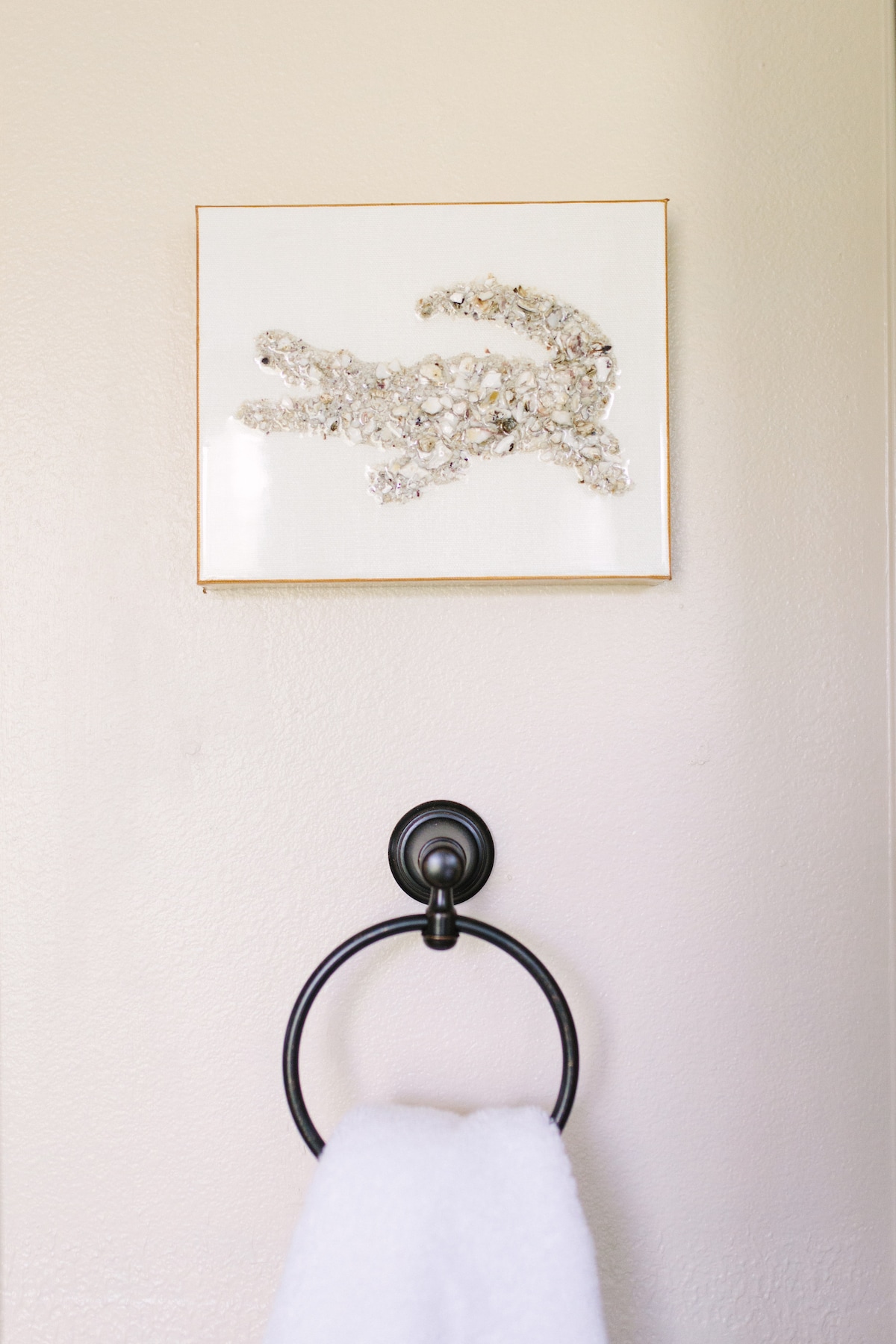 A decorative wall art piece is displayed in a thin golden frame, featuring an abstract design with textured elements. Below, a towel hanging on a black circular towel ring adds a simple, functional touch to the space.