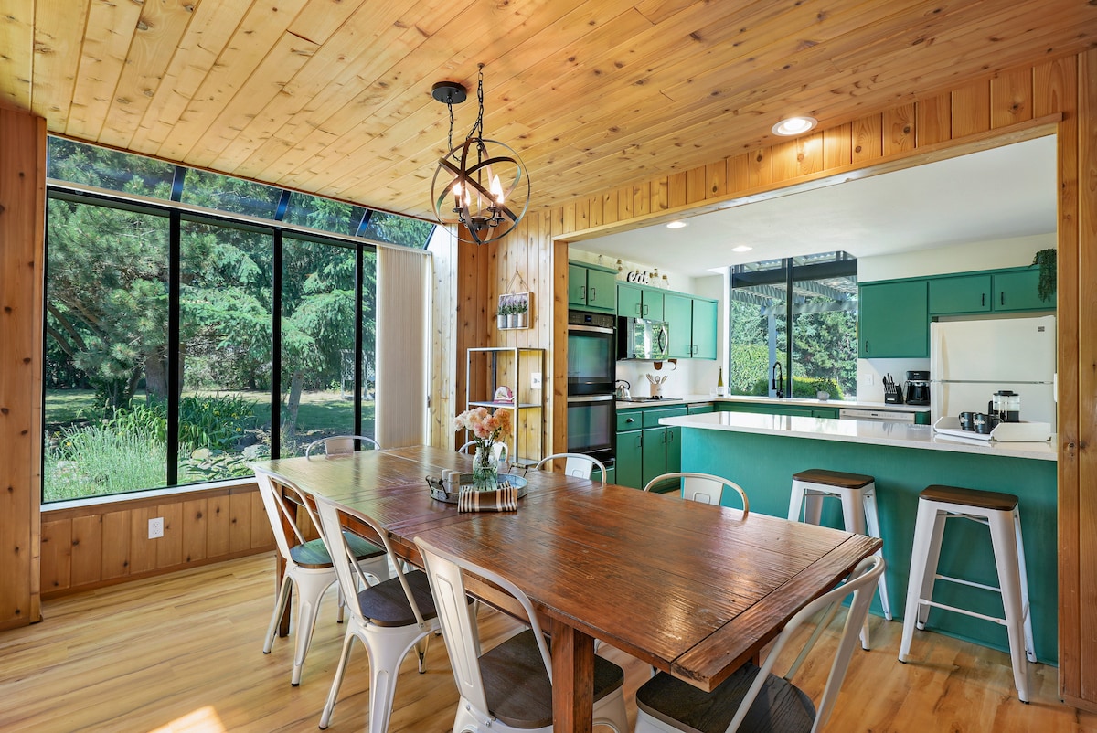 A dining area features a long wooden table surrounded by metal and cushioned chairs. Large windows provide views of the surrounding greenery. The adjoining kitchen is visible, showcasing modern appliances and a casual seating area with bar stools.
