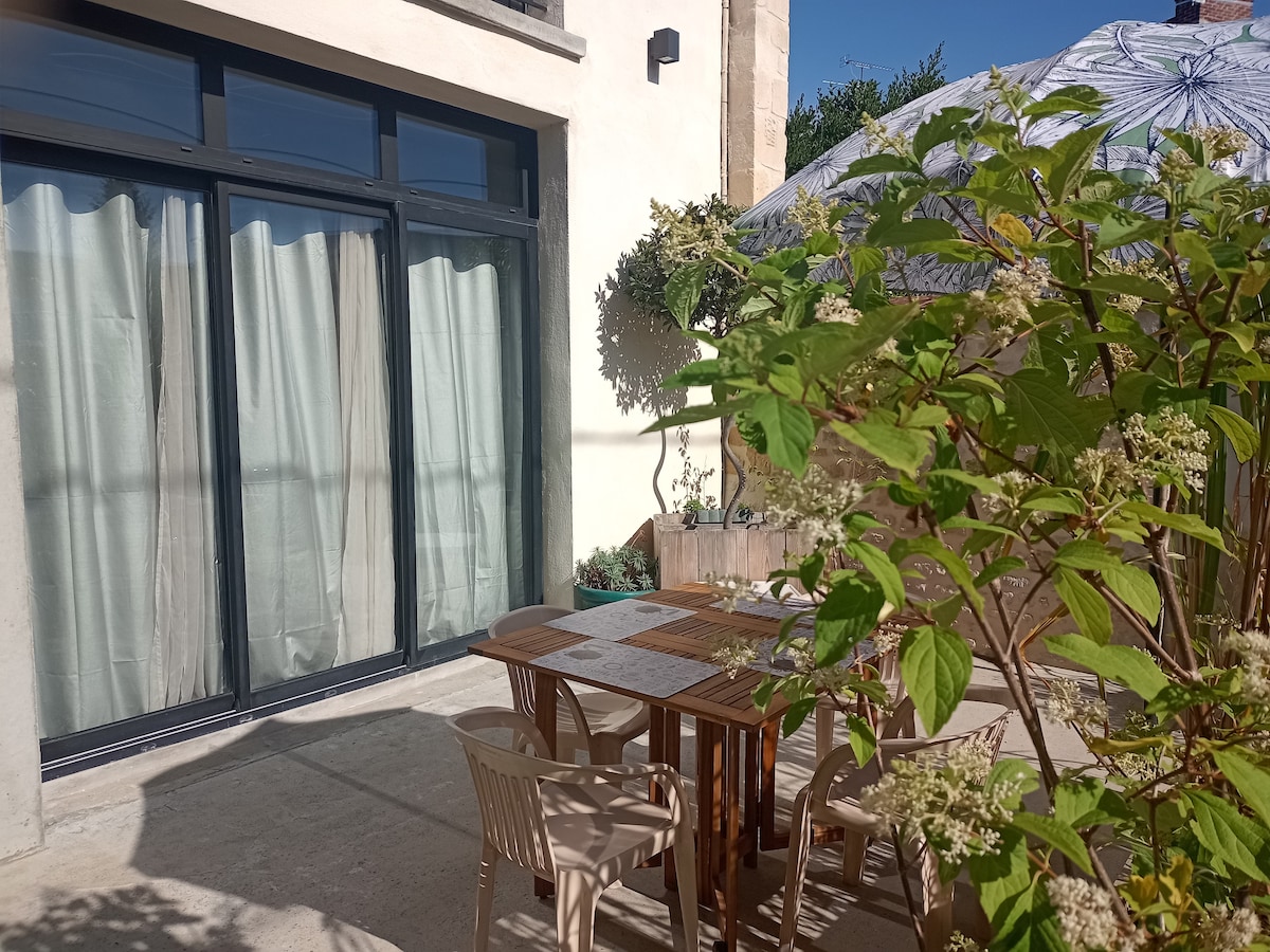 A private terrace is depicted, featuring a solid wooden dining table surrounded by light-colored chairs. Sunlight filters through a large umbrella, casting soft shadows. Lush greenery is present along the edges, enhancing the outdoor setting.