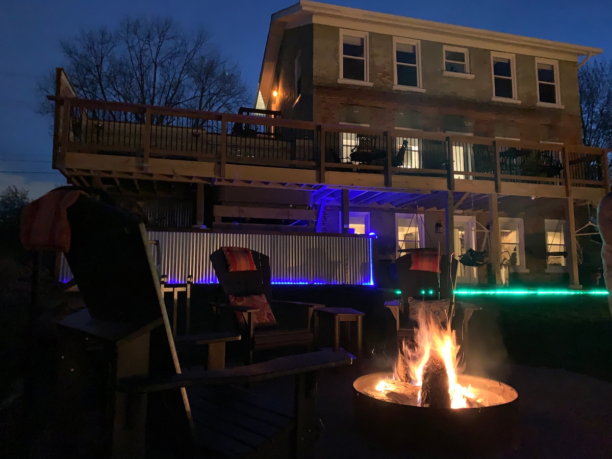 The exterior of the home is illuminated at night, showcasing a spacious deck and a fire pit surrounded by several chairs. Soft lighting accents the structure and outdoor areas, enhancing the inviting atmosphere of the backyard.