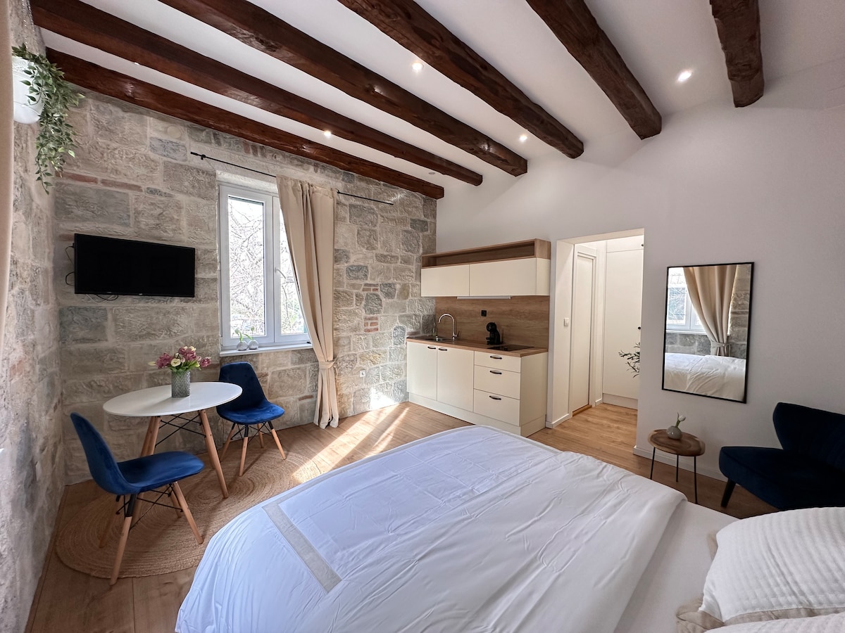 A bright studio space features rustic stone walls and wooden beams overhead. A compact kitchenette is visible, equipped with essential appliances. A small dining table with two chairs occupies one corner, while a spacious bed with white linens dominates the room. Natural light streams through the window.