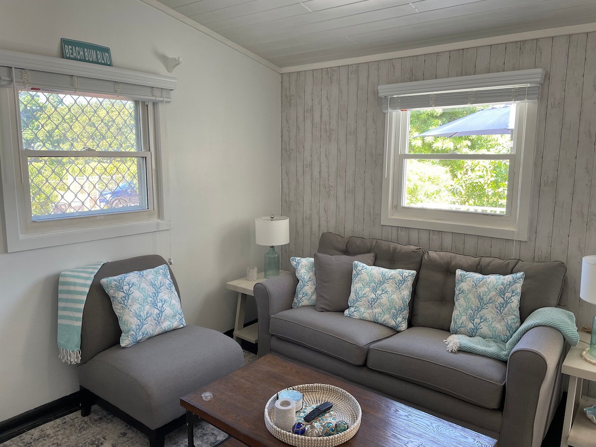 A comfortable living area features a gray sofa adorned with decorative cushions, complemented by a matching chaise lounge. A wooden coffee table holds decorative items, while natural light filters through window screens, creating a bright and inviting ambiance.