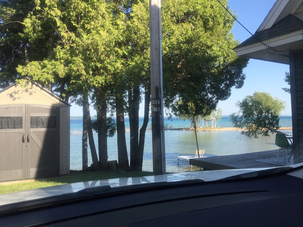 A view of Lake Michigan Bay is framed by lush trees, providing a natural backdrop to the tranquil water. A wooden dock extends from the property, suggesting opportunities for relaxation and outdoor activities.