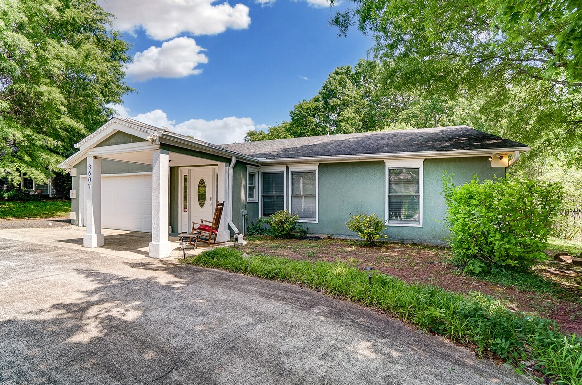 A single-family home is depicted with light blue exterior walls and a welcoming porch featuring a rocking chair. A large driveway leads to a garage, surrounded by greenery and large trees that provide shade.