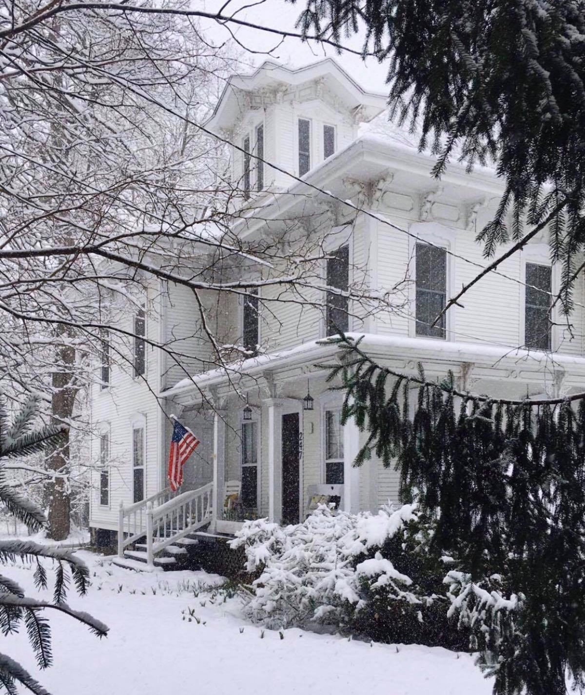 A charming historic mansion is surrounded by a soft blanket of snow, with intricate architectural details visible on the white exterior. An American flag is displayed on the porch, while evergreen trees frame the scene, creating a serene winter atmosphere.