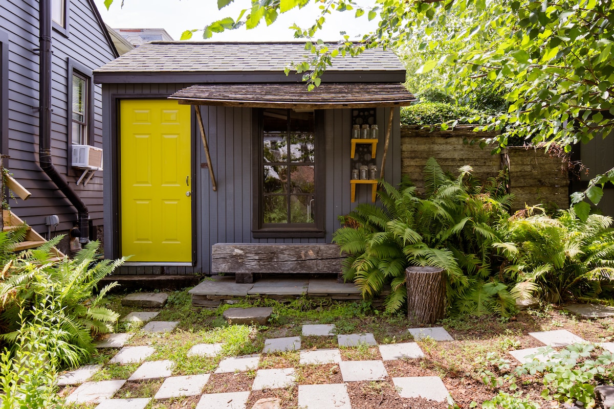 A charming garden shed is seen with a vibrant yellow door, set against a backdrop of lush greenery. The path leading to the entrance features gray stone squares, surrounded by ferns and plants, creating a natural, serene environment.