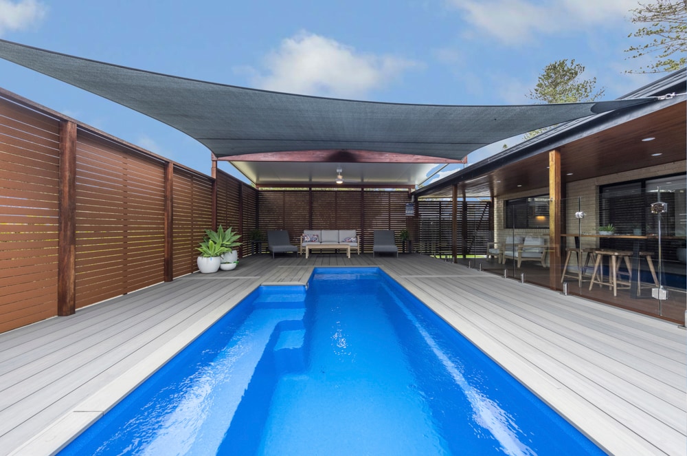 A private outdoor space features a heated plunge pool surrounded by grey decking. A shaded area offers seating with modern loungers and a dining table. Lush greenery adds a touch of nature to the secluded setting.