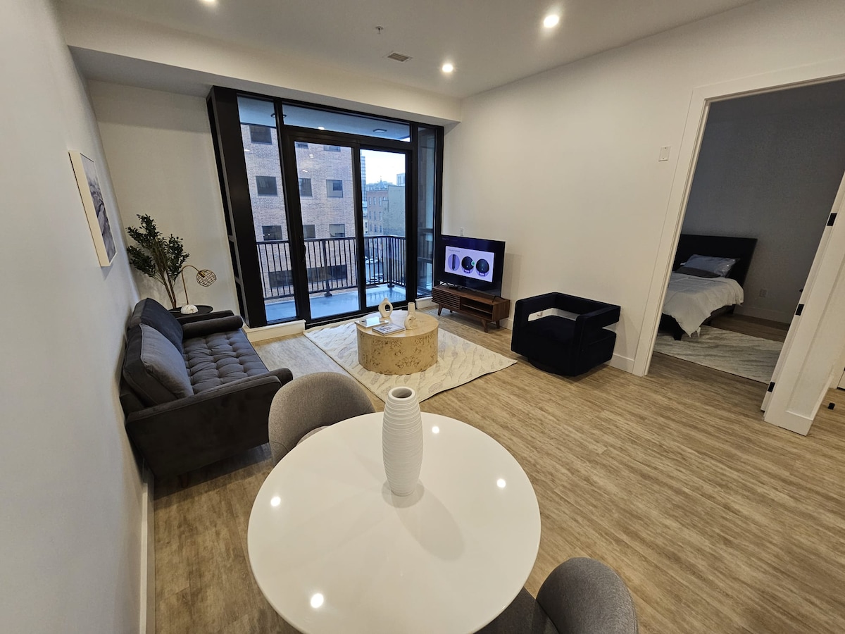 A spacious living area features a plush gray sofa and a modern coffee table, with light streaming in through glass sliding doors leading to a balcony. A round dining table is positioned nearby. A television is visible across from the seating area.