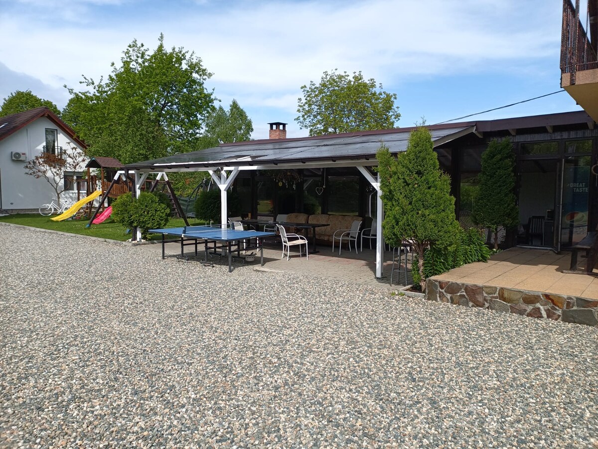 An outdoor area is shown, featuring a covered seating arrangement with a spacious table and several chairs. A ping-pong table is visible, along with recreational elements like swings and slides in the background. Lush greenery surrounds the space, providing a tranquil environment.