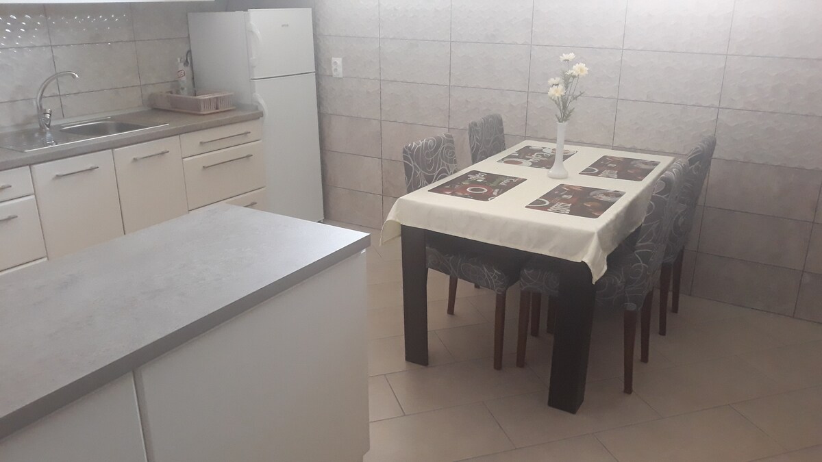 A fully equipped kitchen is visible, featuring modern cabinetry and stainless steel appliances. A dining table is set for six, with a light-colored tablecloth and decorative flowers as centerpieces, surrounded by patterned chairs. Natural light illuminates the tiled space.