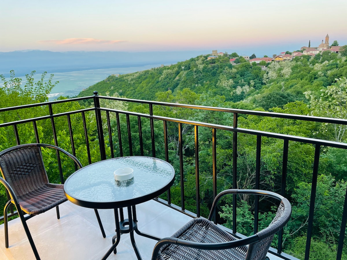A balcony is furnished with a glass-top table and two woven chairs, offering a tranquil space to enjoy the expansive views of lush greenery and distant hills. The horizon features soft pastels as the sun sets, enhancing the serene atmosphere.