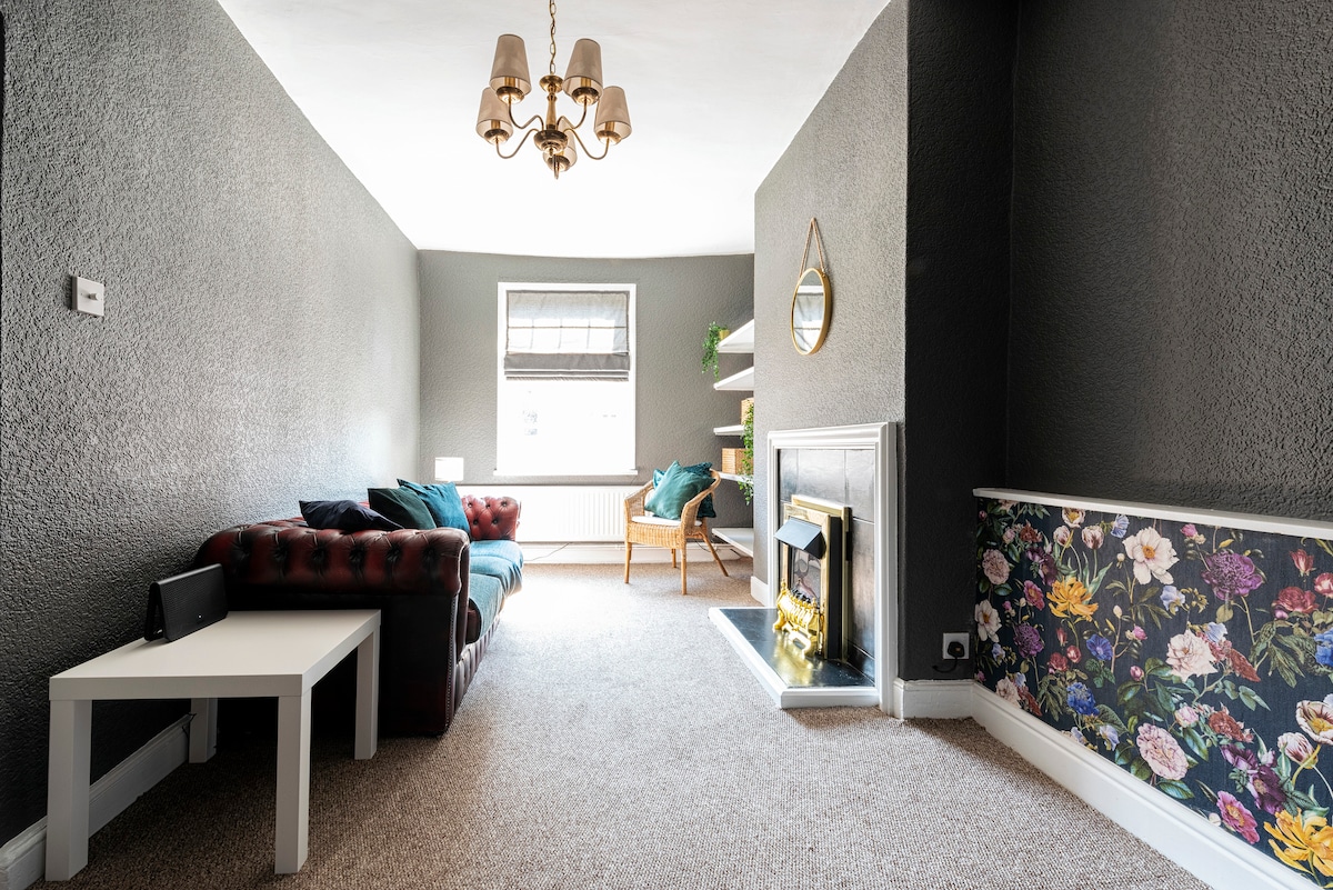 The cozy living room features a dark, textured wall with contrasting floral wallpaper. A deep burgundy leather sofa and a wooden chair create a relaxed seating area. Light enters through a window, illuminating a small side table and a fireplace adorned with a mirror above.
