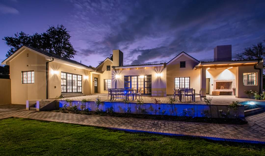 An elegantly designed manor house is illuminated at dusk, with warm lights highlighting the architecture. A spacious patio area features outdoor seating and a tranquil pool reflecting the sky, while well-maintained gardens frame the space.
