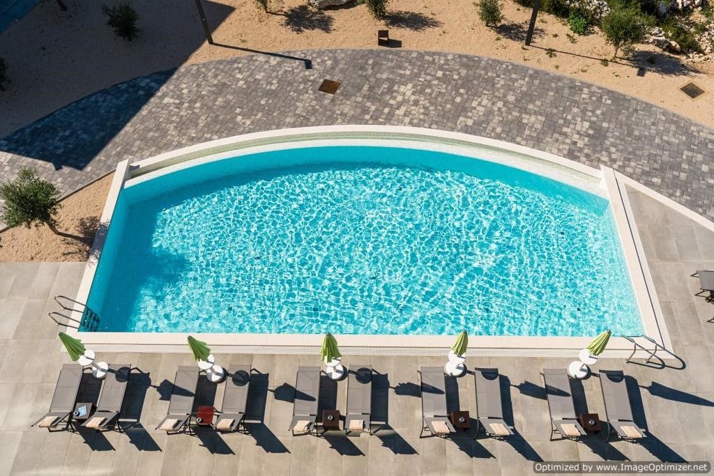 An aerial view of a rectangular pool featuring clear, blue water. Lounge chairs arranged around the pool are complemented by green umbrellas. The surrounding area showcases a stone patio with a minimalist design, bordered by sandy spaces and greenery.