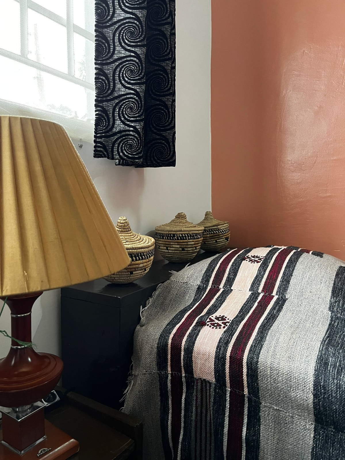 A corner of a room is highlighted, featuring a lamp with a golden shade and decorative woven baskets. A striped blanket is elegantly draped over a bedside table, while a textured wall in warm shades completes the inviting atmosphere.